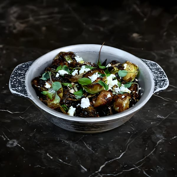 A metal bowl of roasted Brussels sprouts topped with feta cheese and fresh herbs on a dark marble surface.