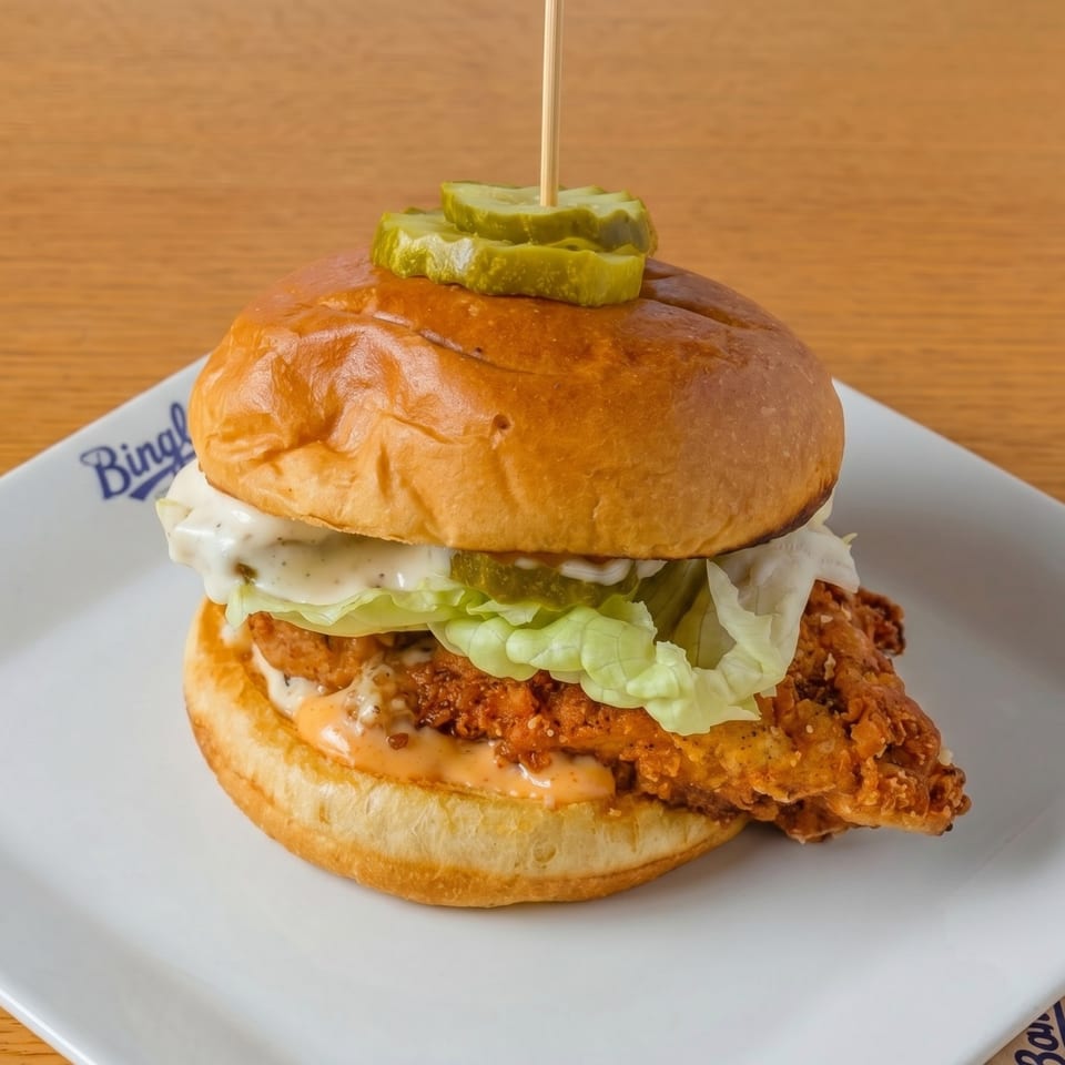 Crispy fried chicken sandwich with lettuce, sauce, and pickles on a brioche bun, on a white plate.