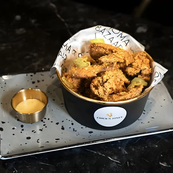 A bucket of crispy Chick N Jones fried chicken wings with pickles and a side of sauce.