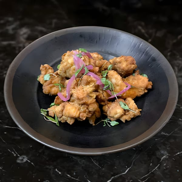 Crispy fried chicken pieces, garnished with pickled red onions and microgreens, served in a dark bowl.